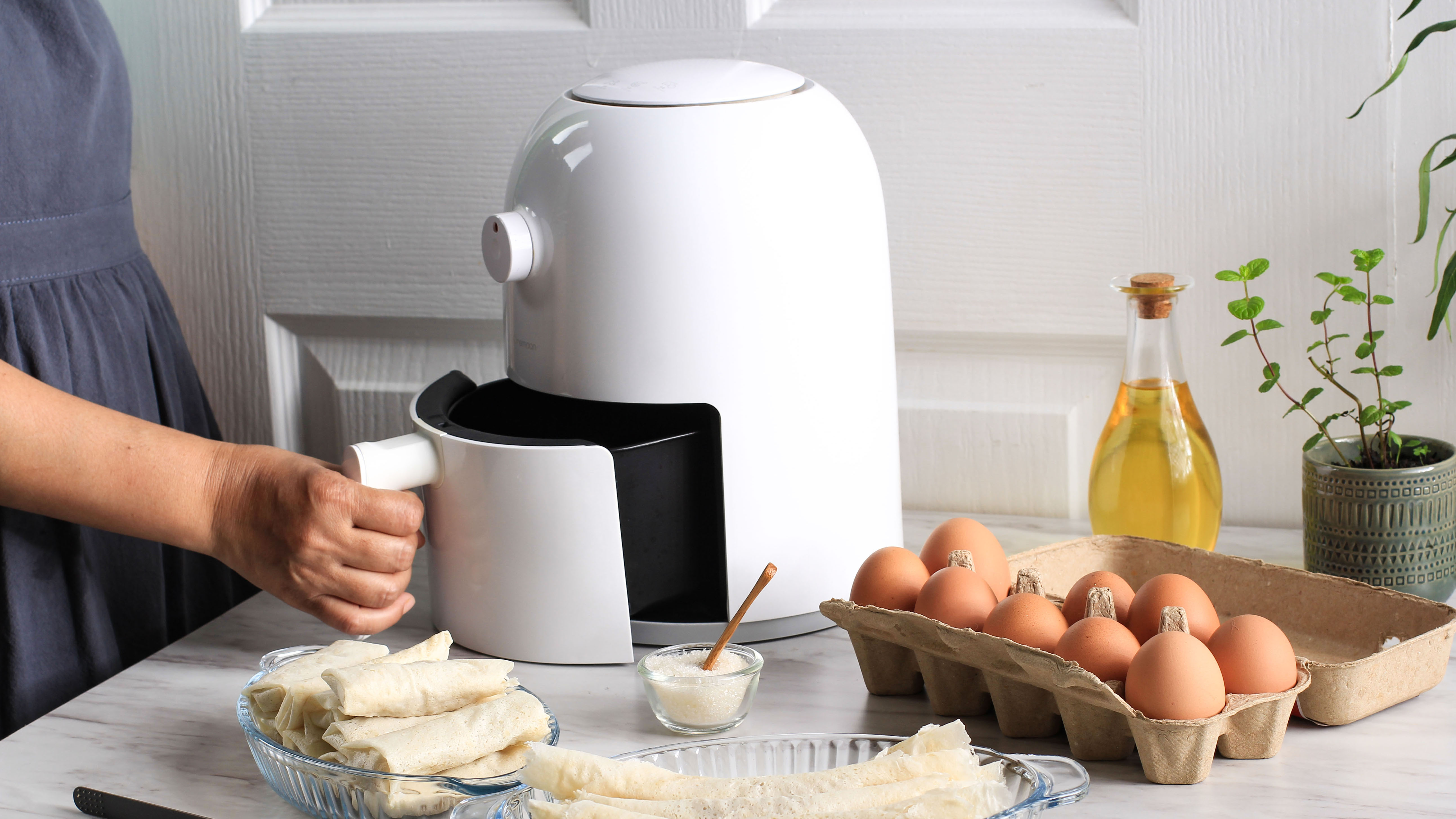 A white air fryer with the basket opened by hand on a table with surrounding ingredients, including eggs and oil