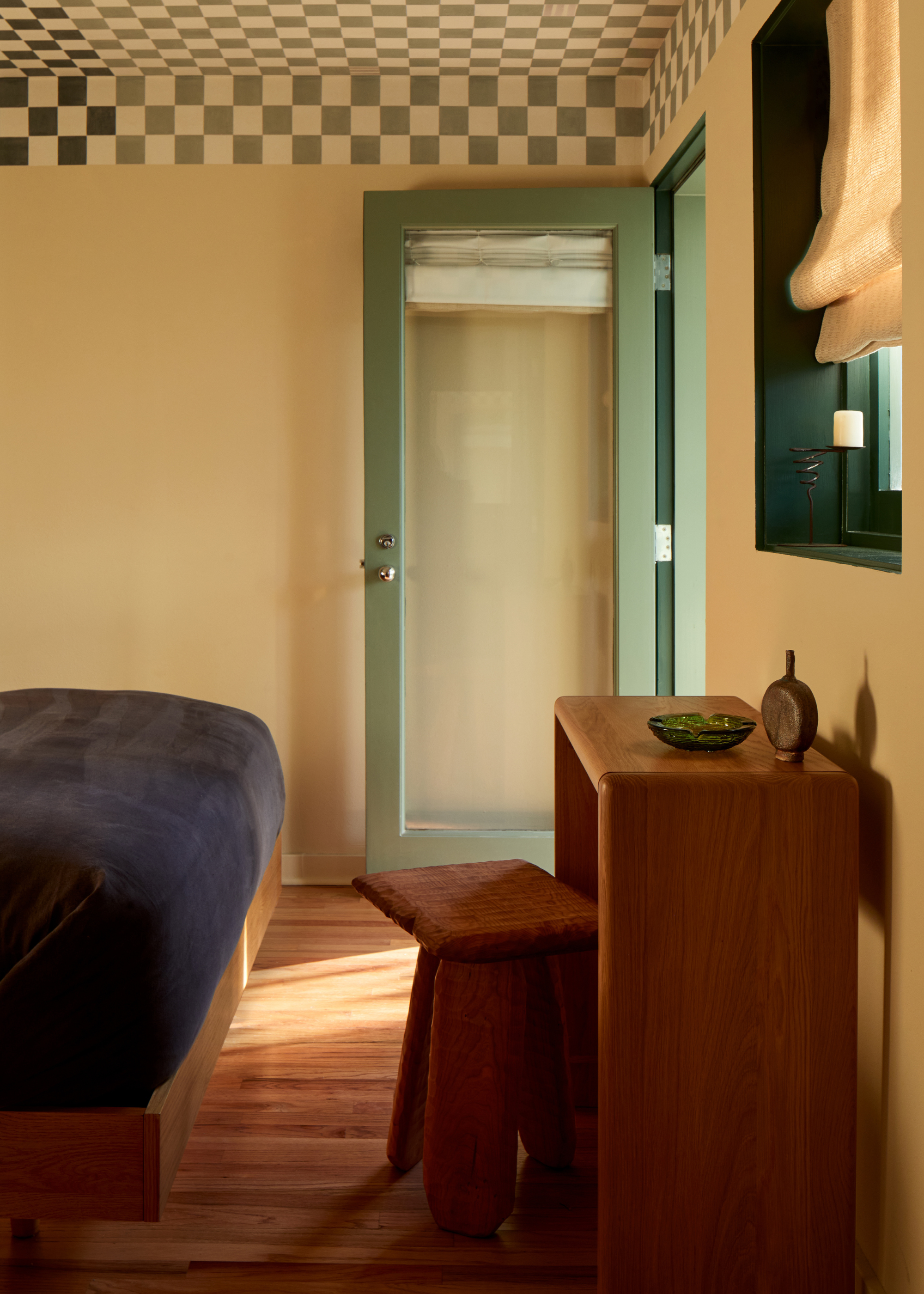 A bedroom with a checkerboard ceiling, a sage green door, a blue bedscape, a wood desk and stool, and a green accent window