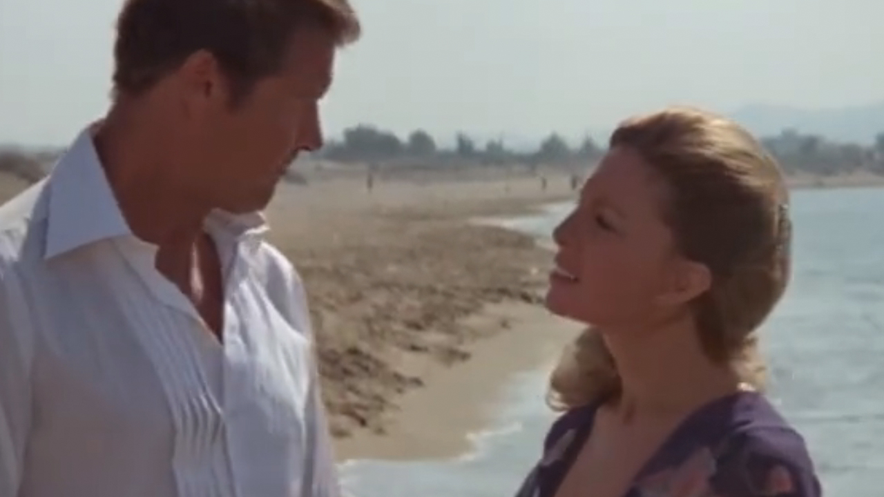 Cassandra Harris smiling and talking to Roger Moore on a beach in For Your Eyes Only