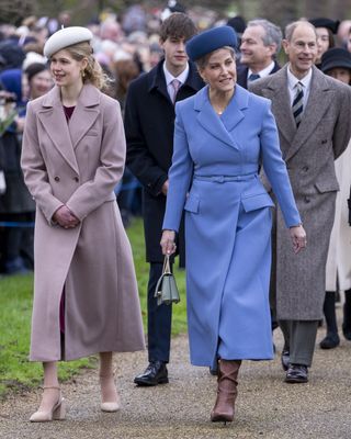 Duchess Sophie wearing a long blue coat and hat walking outside next to Lady Louise wearing a pink coat, with Prince Edward and James walking behind them