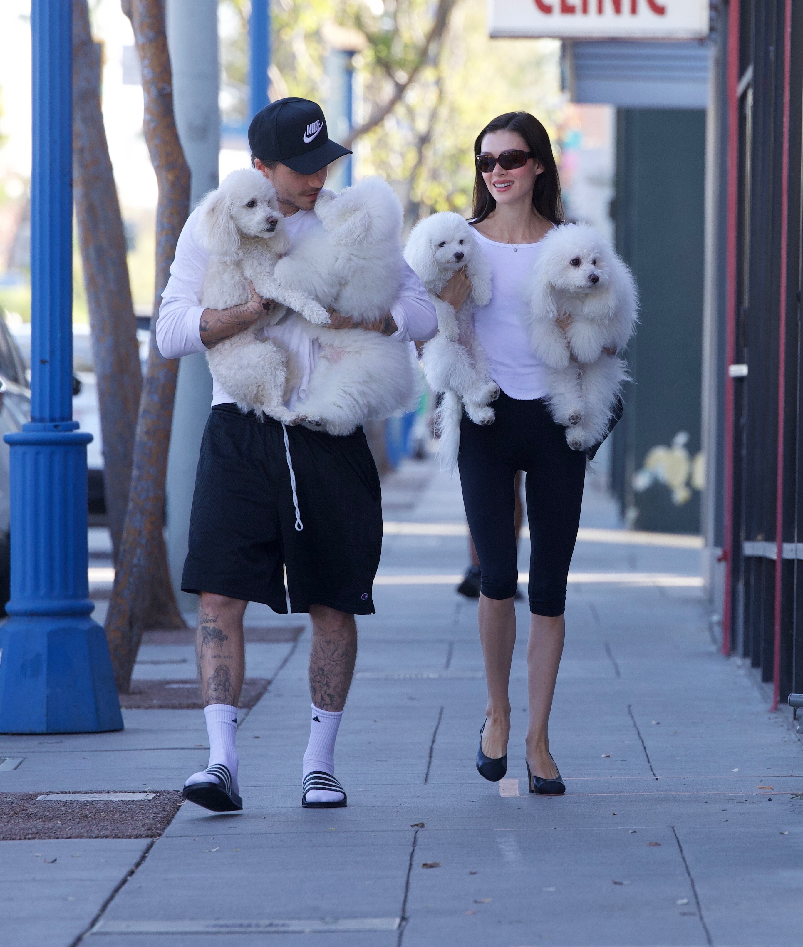 Nicola Peltz Beckham walks alongside Brooklyn Beckham carrying puppies. She wears a white t-shirt with black capri leggings and black rounded-toe heels.