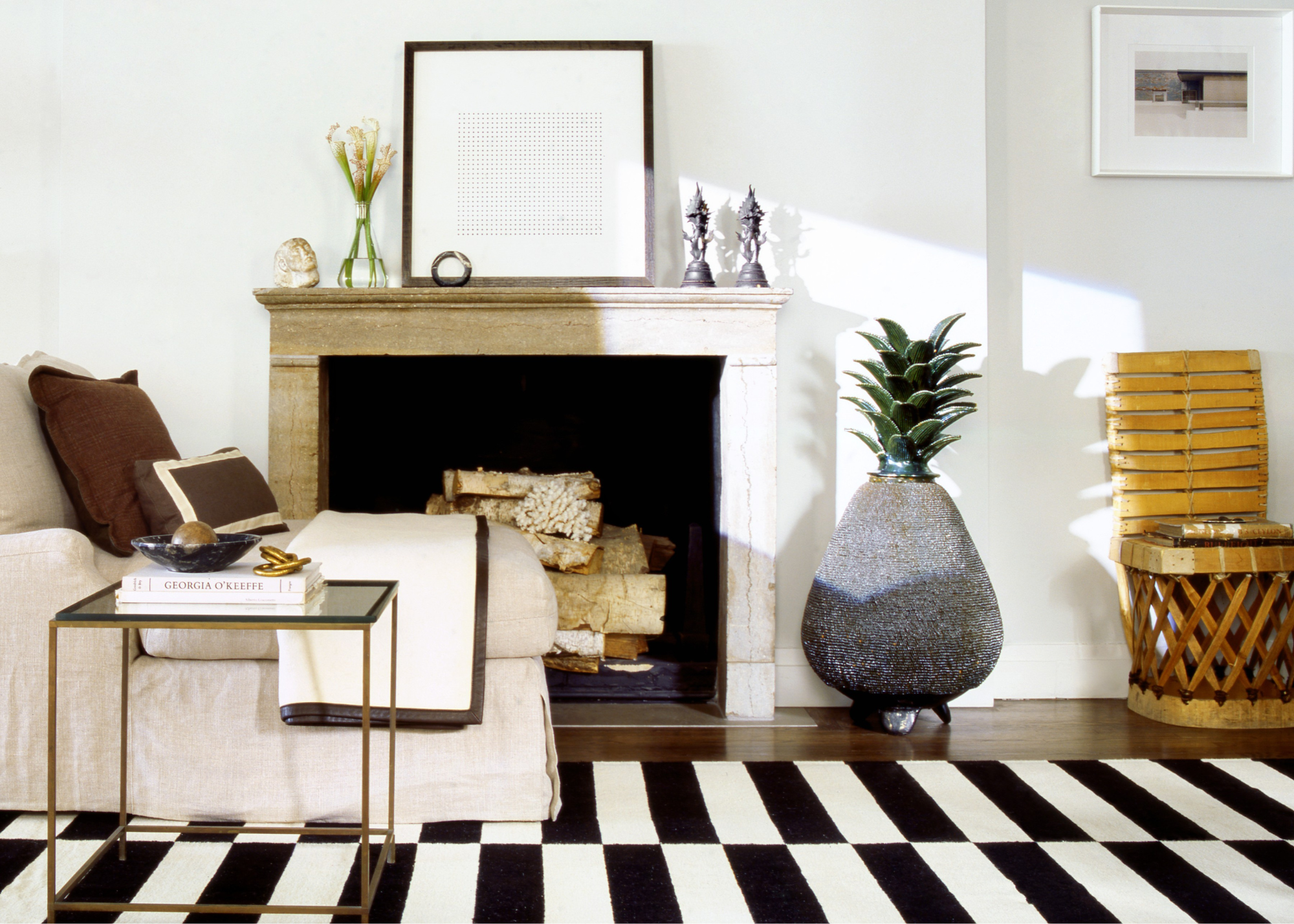 A fireplace with a framed picture resting on top and some decorative items and a vase of flowers, with a black and white checkered rug in front with a cream armchair and side table on top; a large grey vase with a plant inside sits next to the fireplace next to a statement wooden chair