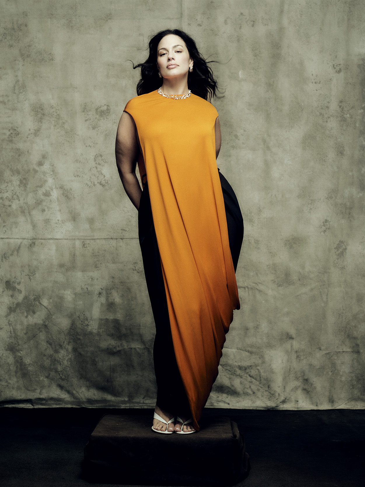 Ashley Graham poses against a grey backdrop wearing a long orange gown and jewels