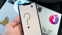 A hand holding an iPhone displaying a black hand-drawn question mark in the Microsoft Whiteboard app, resting over a sticker-covered laptop.