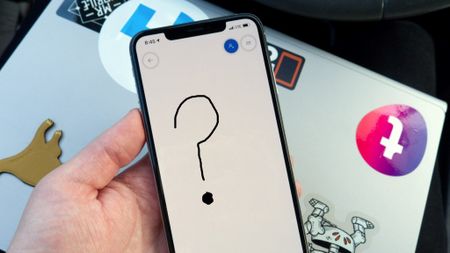 A hand holding an iPhone displaying a black hand-drawn question mark in the Microsoft Whiteboard app, resting over a sticker-covered laptop.