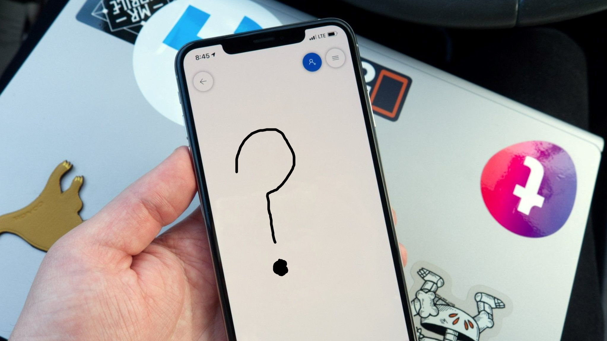 A hand holding an iPhone displaying a black hand-drawn question mark in the Microsoft Whiteboard app, resting over a sticker-covered laptop.