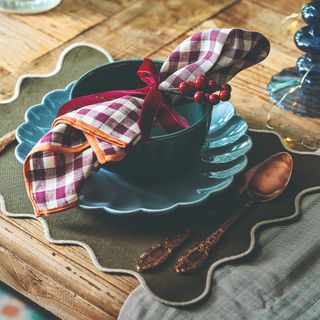 A rustic wooden dining table set for a Christmas dinner with a scalloped green placemat and gingham napkins tied with a red velvet ribbon