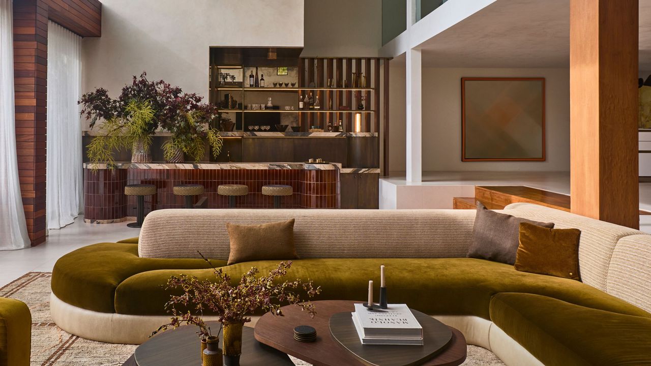 modern living room with curved custom velvet two-tone sofa, tiled home bar with large floral display and backlit shelves, timber panelling, a nesting coffee table, and step up to a higher room with timber beam and polished concrete finishes
