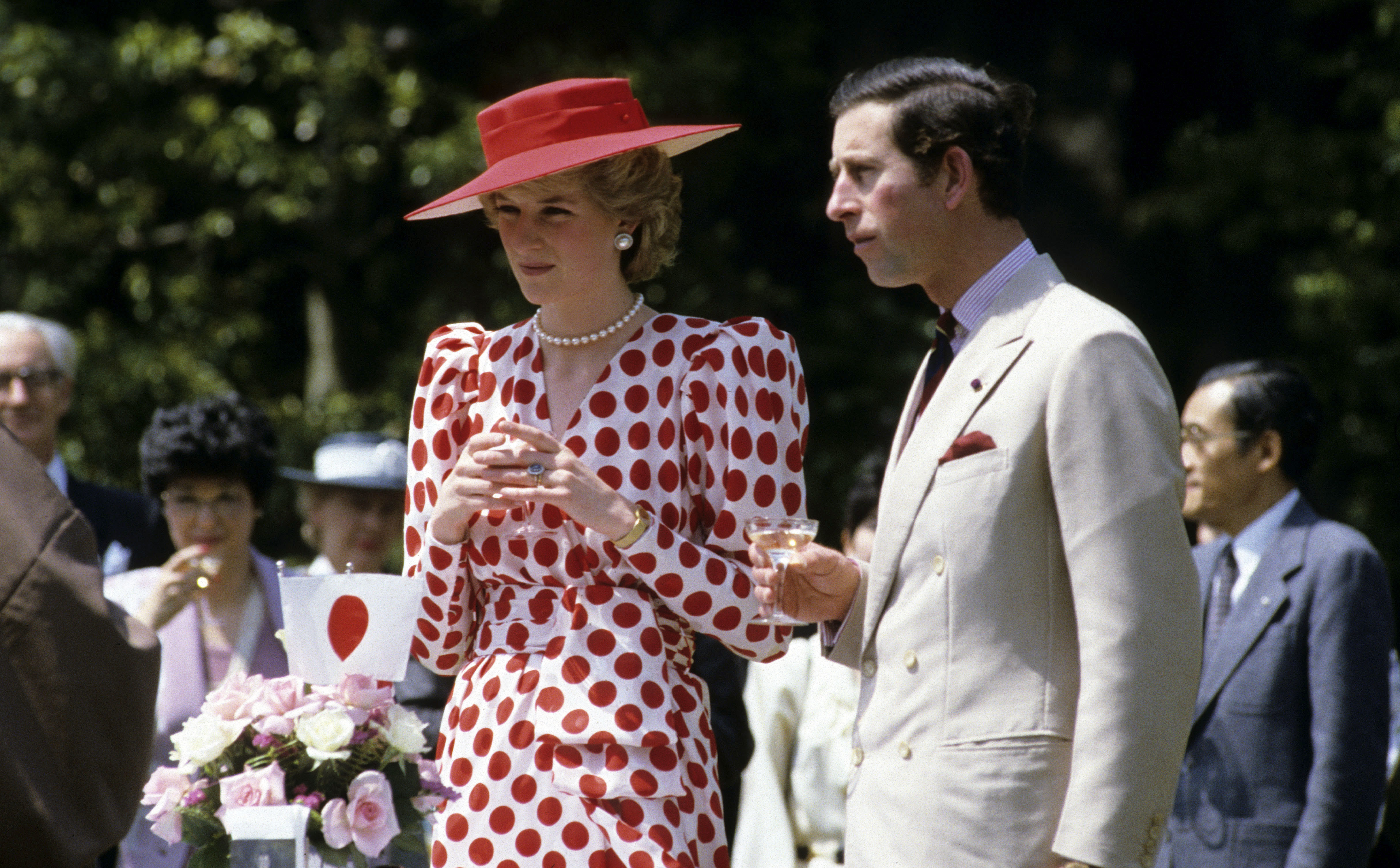 Princess Diana wearing a red polka dot dress and red hat standing next to Prince Charles