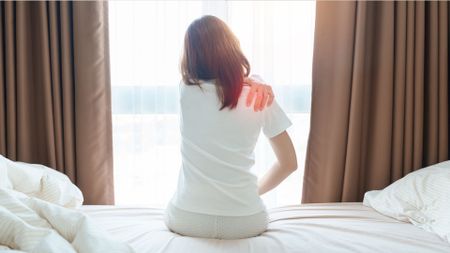 The image shows a woman sitting up in bed and clutching her shoulder in pain