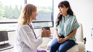 Female patient is shown sat on a white chair at the doctor&#039;s office. She has her hands crossed on top of her lap and is looking at a female doctor who appears to be explaining something to her. 