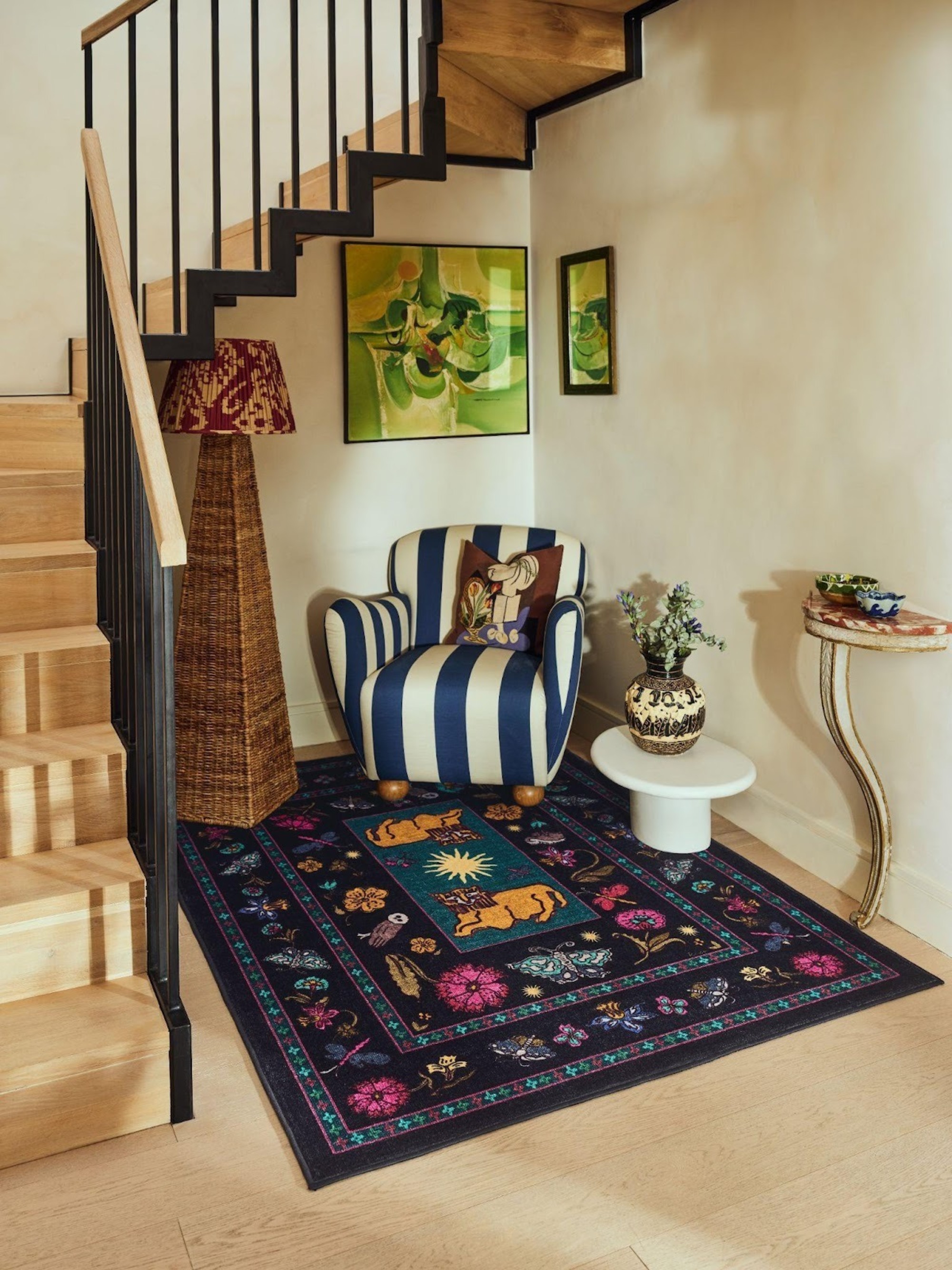 Image of a stairwell/entryway with a seating area under the stairs. There is a blue and white striped chair, a jute floor lamp, a small white side table, and a navy rug.