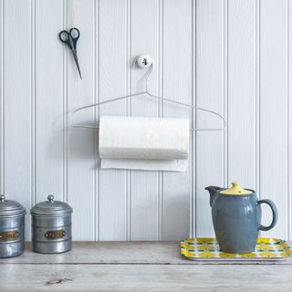 Wire clothes hanger hanging on a wall with a kitchen roll hanging from it, next to sugar pots and a teapot