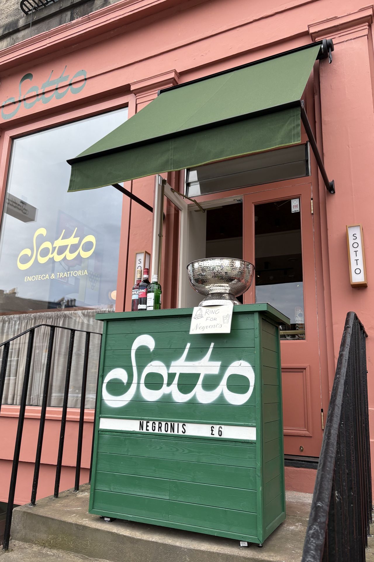 A terracotta-painted restaurant facade features a green awning with matching outdoor bar in wood that reads "Sotto" in white and "Negronis &pound;6" in black on white background.