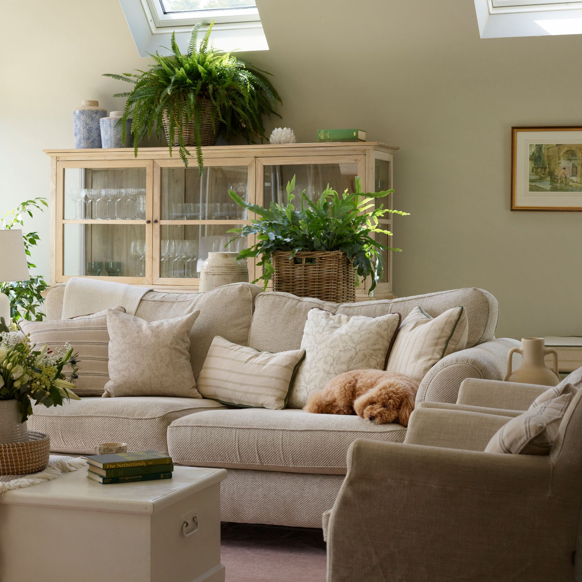 Beige living room with neutral sofa filled with cushions, cream wood trunk coffee table and pine wood cabinet filled with glassware, lots of greenery