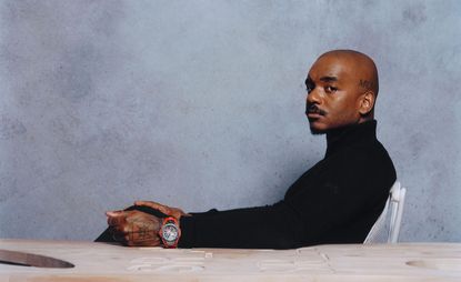 Samuel Ross sitting in a white chair at a wooden table wearing a black jumper and Hublot watch