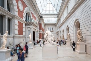 A historical museum with glass ceilings and brick walls houses a series of antique marble statues surrounded by walking visitors.