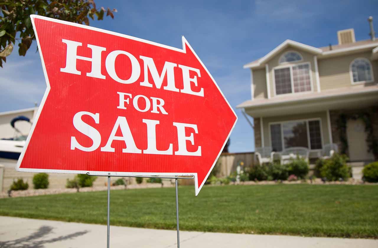 An arrow shaped red &amp;quot;Home For Sale&amp;quot; sign in front of a suburban 2-story home.The green grass and blue sky is visible in the background.