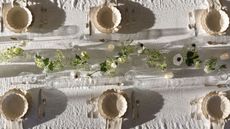 Outdoor tablescape with white linen tablescape, cream scalloped plates and bowls, a floral garland in the centre, and lit candles