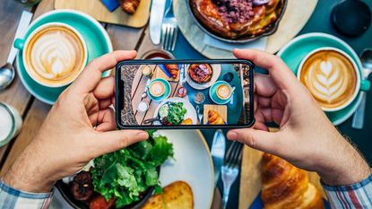 Hands hold a phone taking a photo of a table full of food.