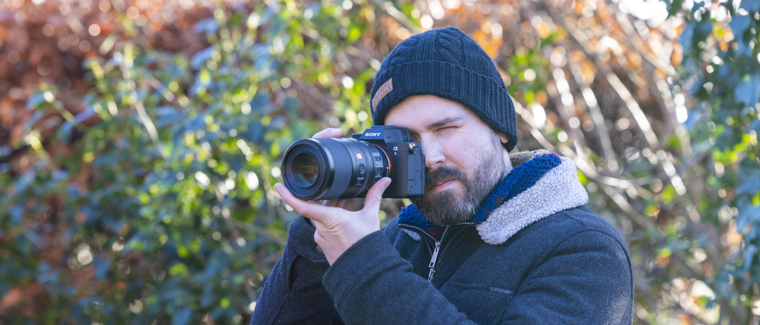 Professional photographer Dan Mold photographed outdoors holding a Sony A7R IV camera to his eye with a Sony FE 100mm f2.8 Macro G Master OSS lens attached to it