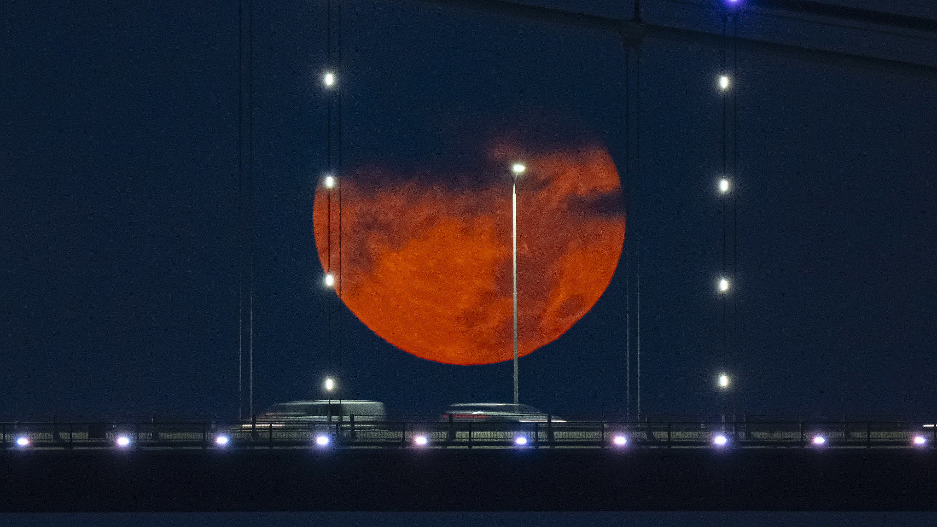 A red full moon is pictured between the supports of a suspension bridge against a night sky, with the upper part of its disk shrouded in cloud. The blurred forms of moving cars can be seen travelling along the bridge and bright lights are placed along the bridge and suspension supports.