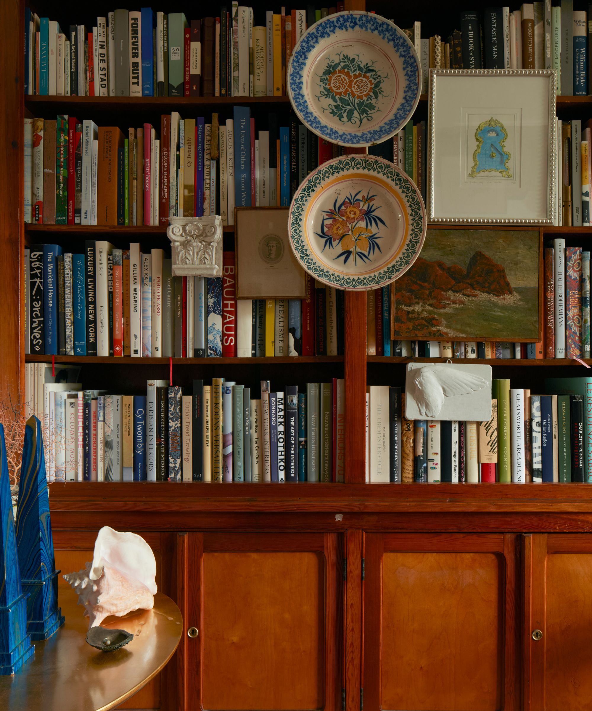 Dark wood bookcase with lots of books and ceramic plates hung in front, and vintage paintings and prints beside a gold round table with a shell object and paperweights