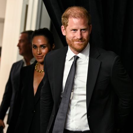 Prince Harry wears a black suit and has red hair and is holding hands with wife Meghan Markle who is wearing a black shirt dress