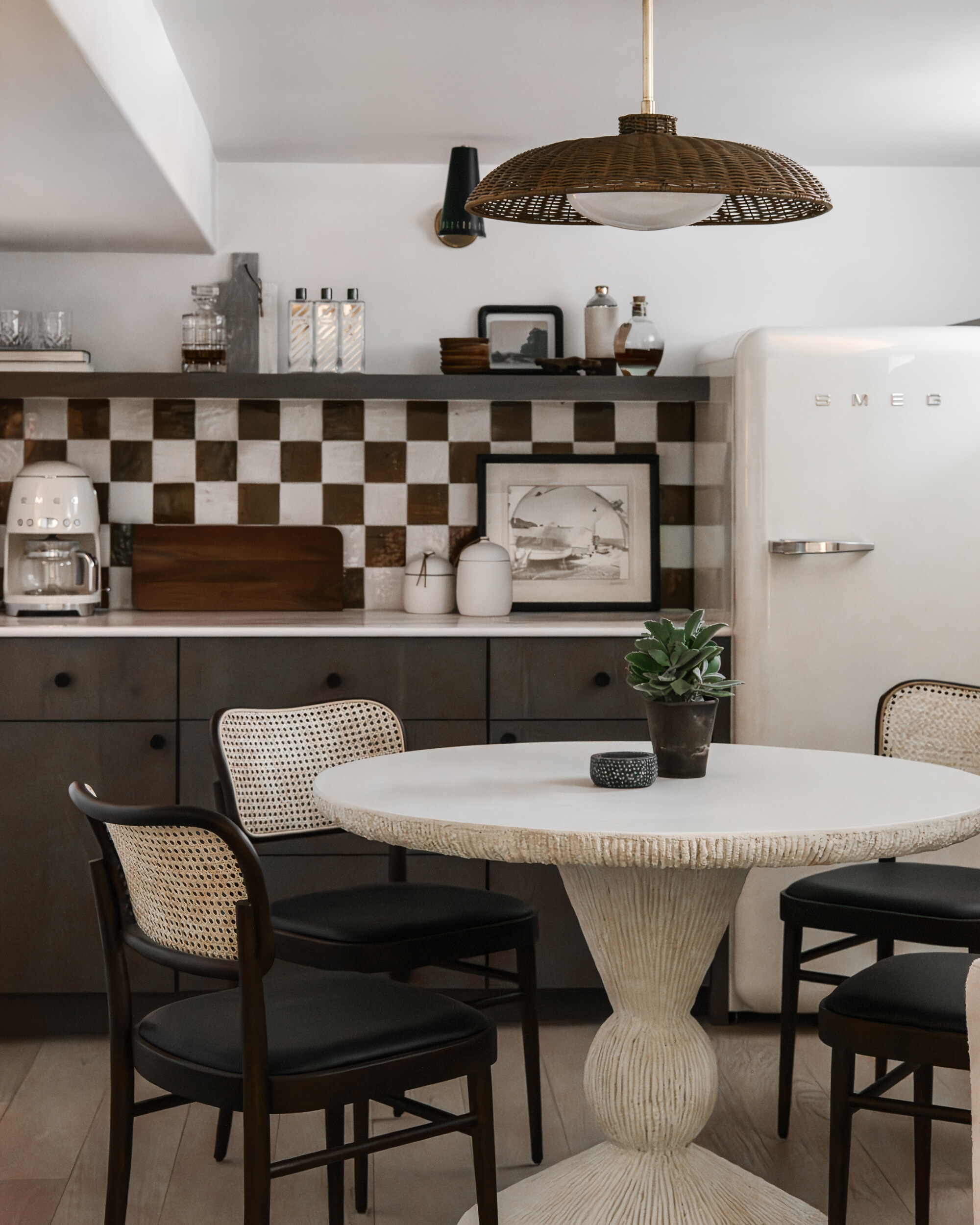 Image of a white and brown kitchen with a checkerboard backsplash and a round, white dining table in the center. There is a white Smeg fridge and a matching Smeg coffee maker on the counter.