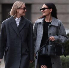Marlies Pia Pfeifhofer (L) seen wearing brown oval sunglasses, silver earrings, white cotton buttoned blouse / shirt, COS dark grey short oversized wool coat, Dior black / white herringbone checked pattern suit vest / matching blazer jacket and suit pants, Dior black / white tweed crossbody bag, Dior black shiny leather loafers; Anna Winter (R) seen wearing Celine black sunglasses, Ganni black fringed short dress, Ganni light grey wool long coat, Loewe black leather handbag and Prada black shiny leather boots, on November 30, 2023 in Berlin, Germany. (Photo by Jeremy Moeller/Getty Images)