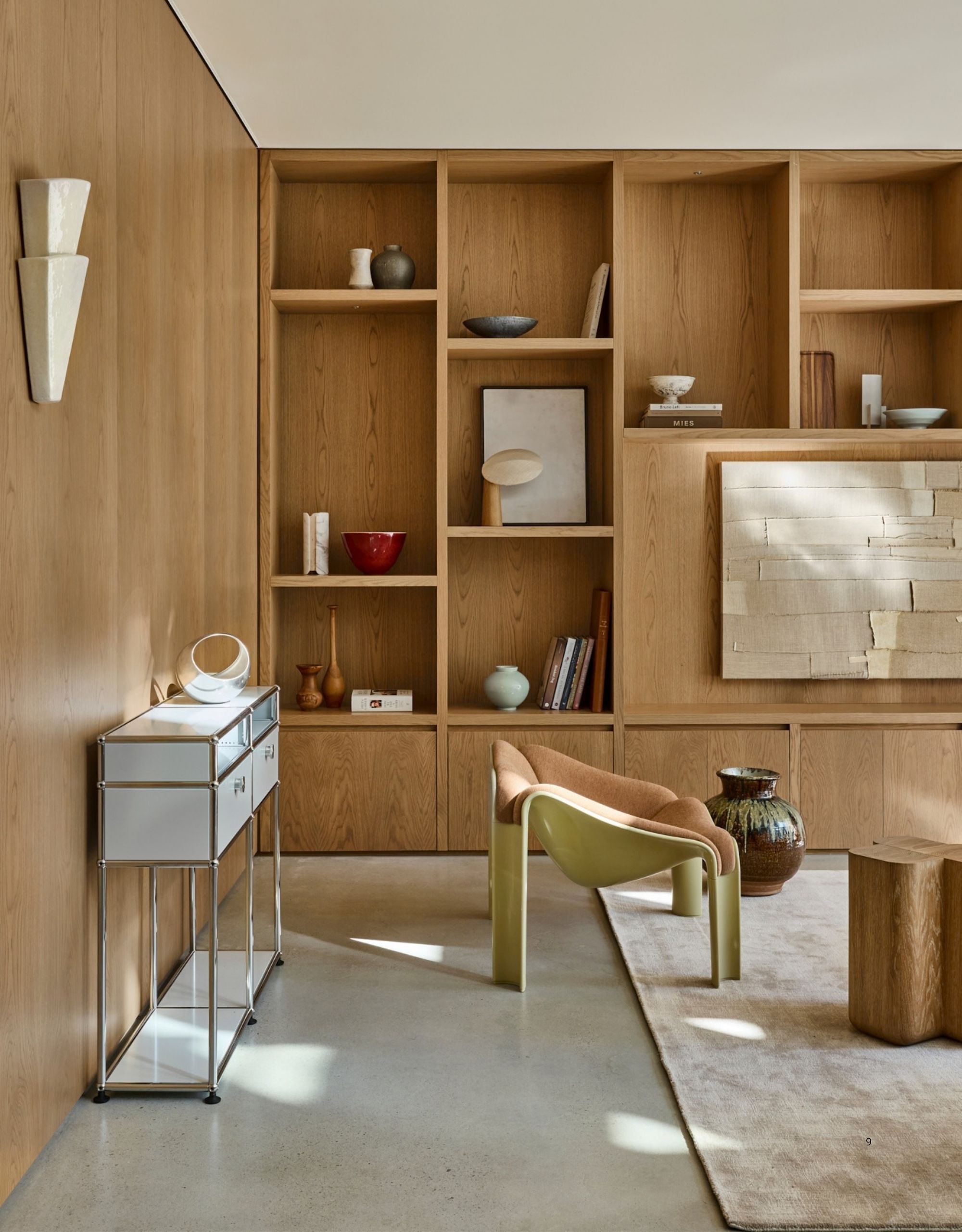 A modern living room with wooden walls and a built-in shelving unit. There are lots of vases, books and art on the shelves. There is an accent chair and a white, metal console in view.