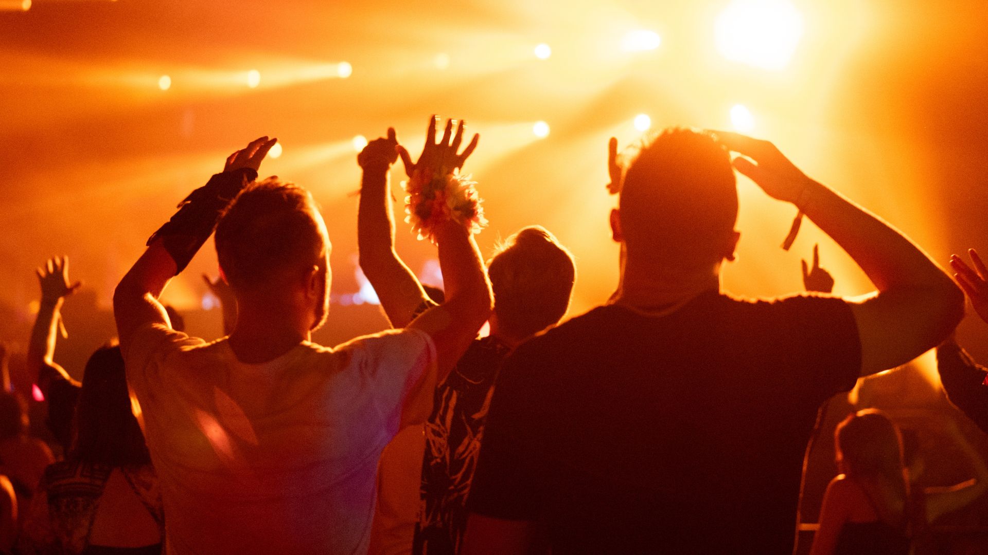 Crowd of people attending a musical performance. Audience dancing illuminated by orange lights - stock photoLarge concert venue filled with a crowd of people. Performers on stage and audience cheering. Strong neon lighting and smoke effects