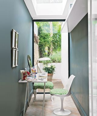 Narrow study area with parquet floor, dark blue grey walls, glass wall to garden small white desk and Eero Saarinen tulip chair with green cushion.