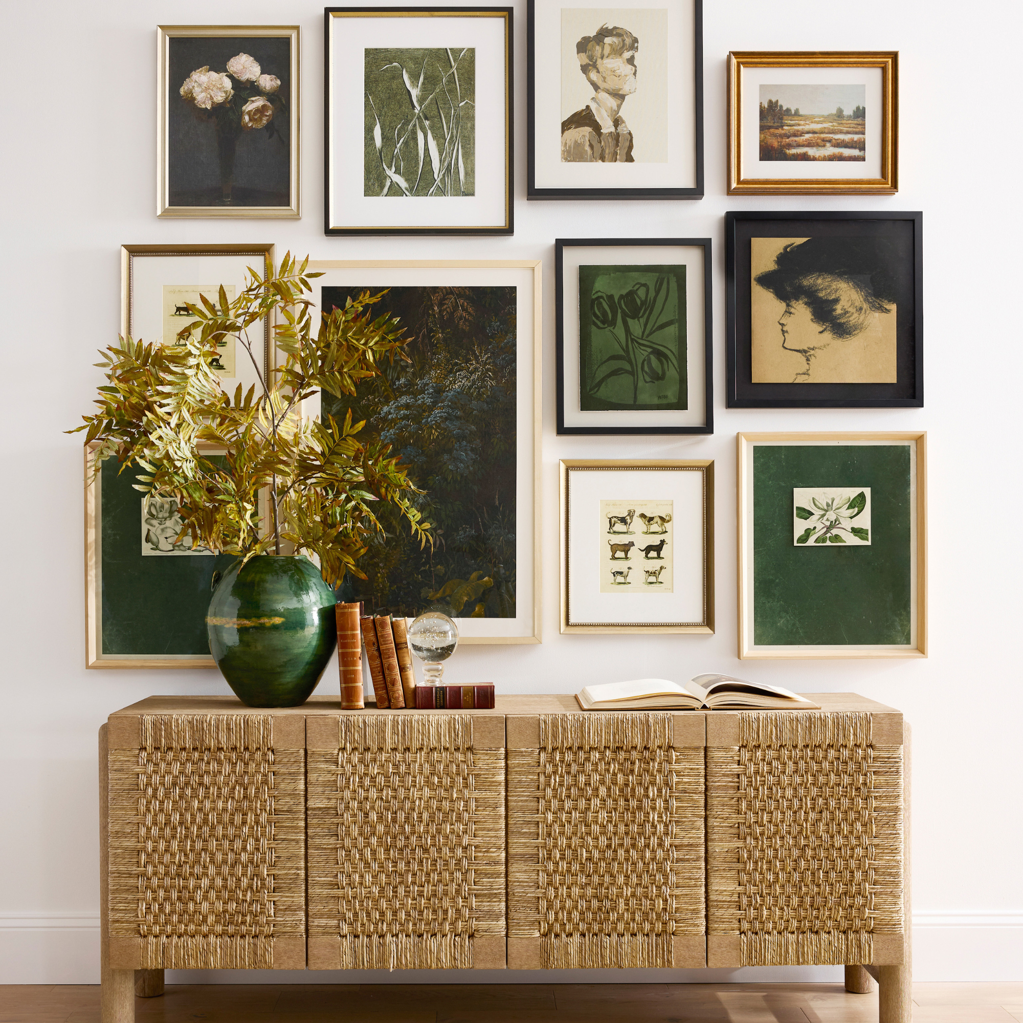 Woven sideboard with books and a green vase on top. Behind is a green and gold gallery wall.