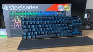 SteelSeries Apex Pro TKL Wireless propped against its packaging with wrist rest on a wooden desk