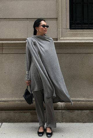 one of the best chunky cuff bracelets is shown in a photo of a woman standing outside wearing a gray cape over a gray sweater with matching knit pants styled with matching black ballet flats, a black bag, black sunglasses, and a silver cuff