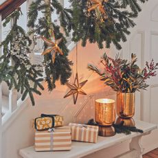 A festive fir garland running up a white banister with metallic stars hanging down and wrapped presents on a wooden bench