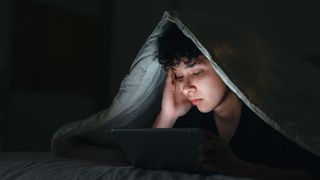 A teen lies in bed in the dark with his face lit up by a bright phone screen