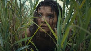 Still from the movie ' Kingdom of the Planet of the Apes' (2024). Close up of a young woman's face hiding amongst the tall, green grass. She looks worried and is covered in dirt.