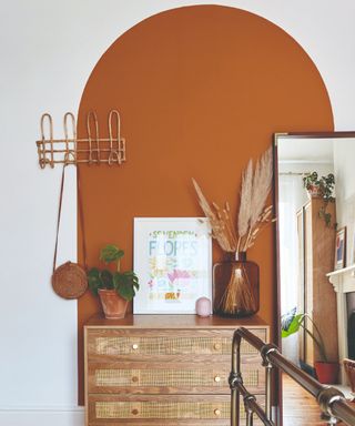 A bedroom with a chest of drawers and a full-length mirror zoned with a large rust-coloured painted arch on the wall