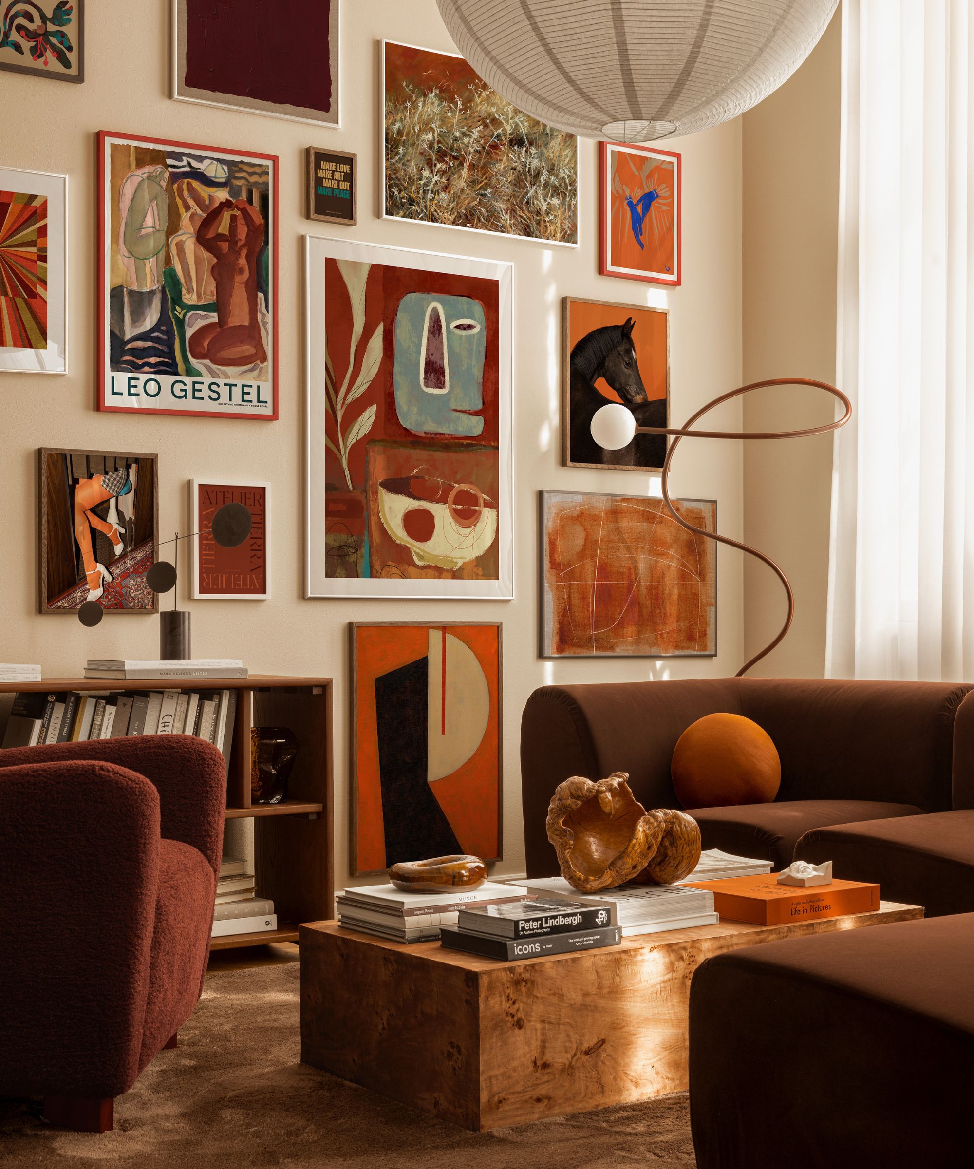 A living room with a gallery wall of framed artwork in warm reds, browns and creams. Soft seating surrounds a low wooden table. Light filters through curtains, creating a cosy, layered space rich in colour, texture and visual interest overall.