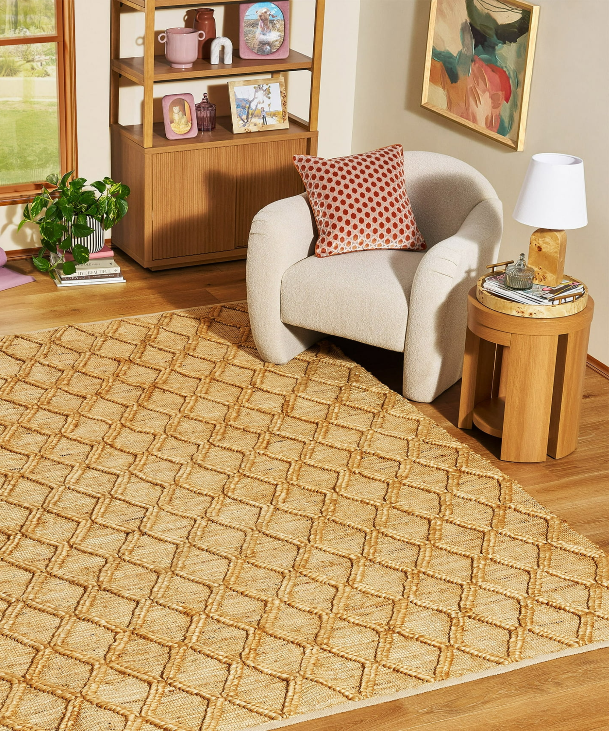 a living room with a jute rug with a trellis pattern, a cream accent chair with a red pillow, a small side table with a table lamp, and a bookshelf in the background holding picture frames and vases