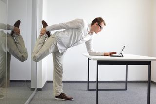 A man stands at his desk, and stretches out his quads as he works.