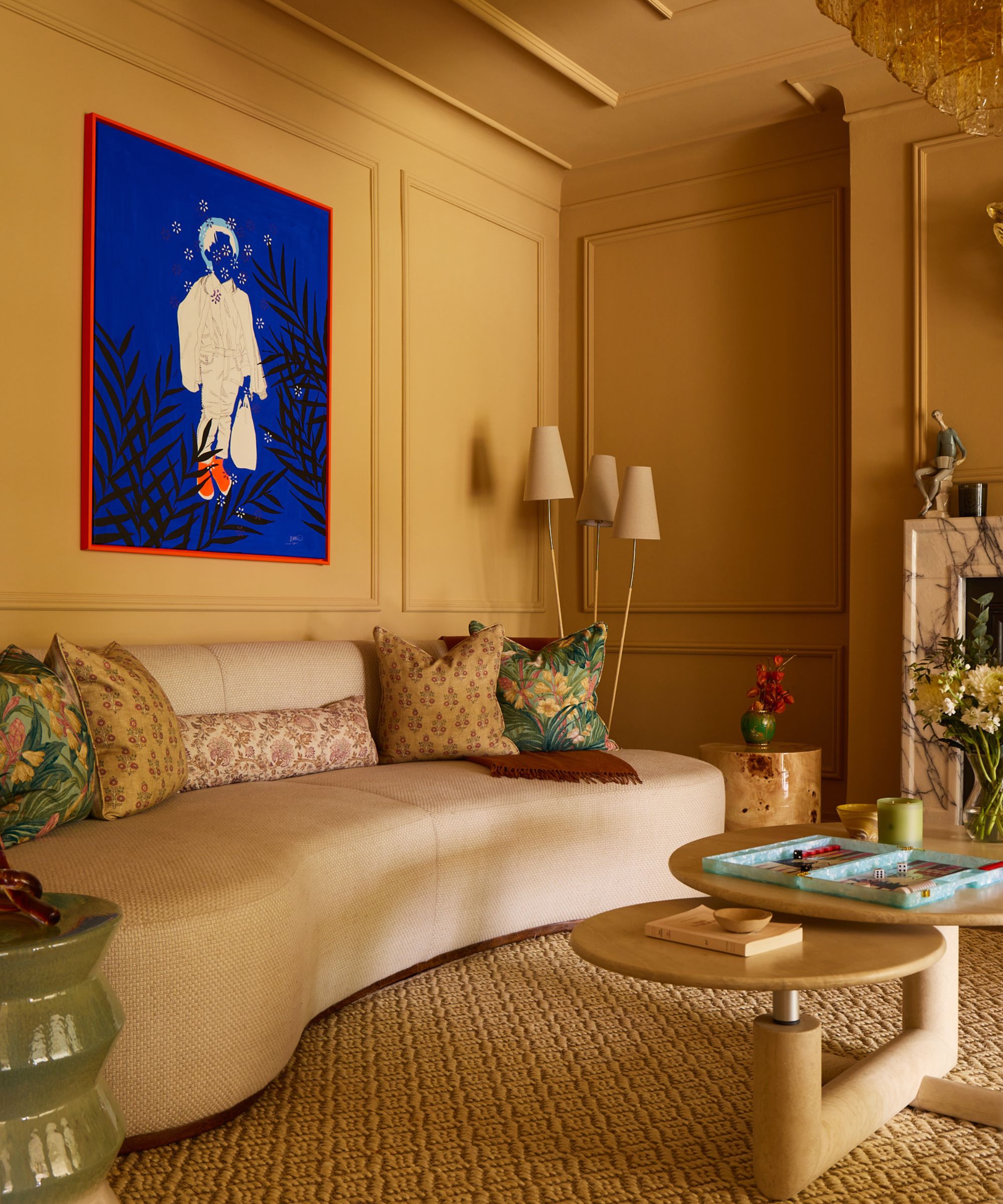 Living room with deep mustard walls, marble fireplace, a large cream curved sofa decorated with block-printed cushions, a round tiered coffee table, and a large cobalt blue painting on the wall.