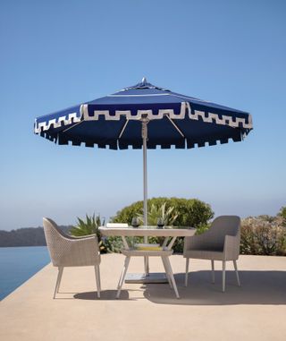 A blue and white parasol with a table and chairs below it with a view of the ocean.