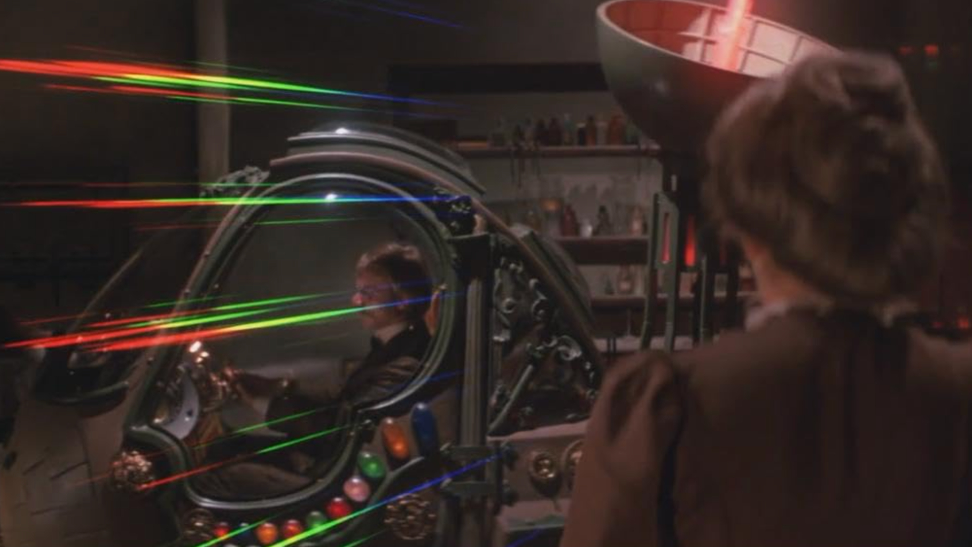 A man is sitting in a car-esque machine with lights flashing around it within a laboratory as a woman watches on