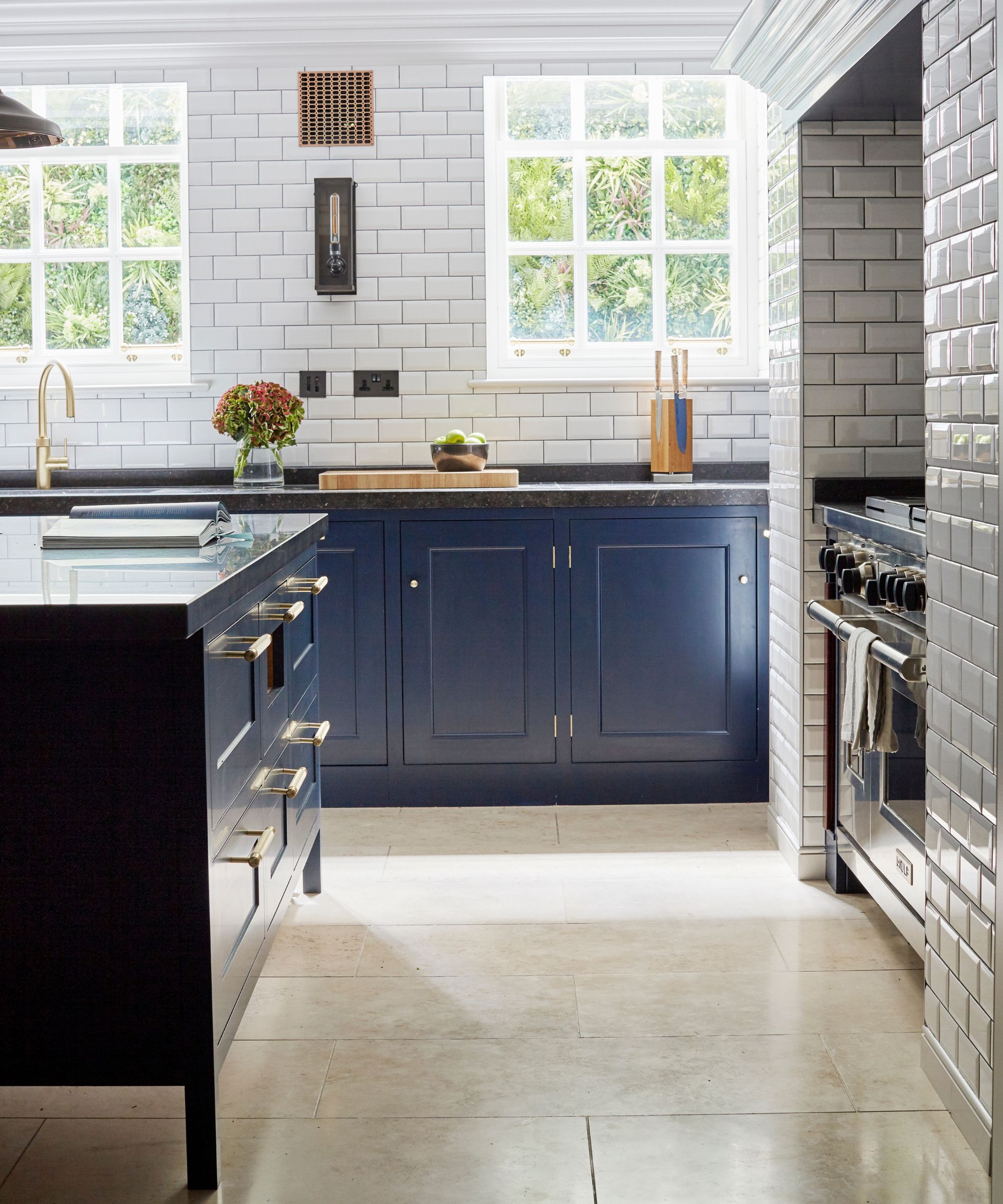 A white tiled kitchen with blue cabinets, gold hardware and faucets, and a brown shiny tiled floor