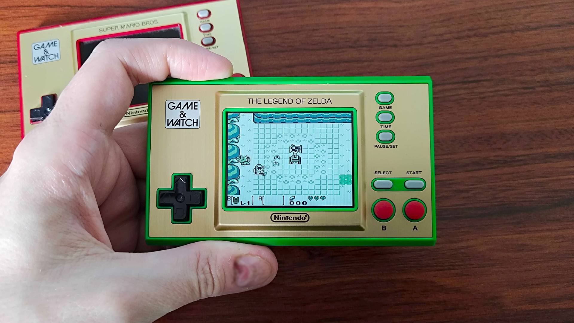 Hand holding Zelda Game & Watch handheld with Link's Awakening on screen and Mario version on woodgrain desk at top left corner.
