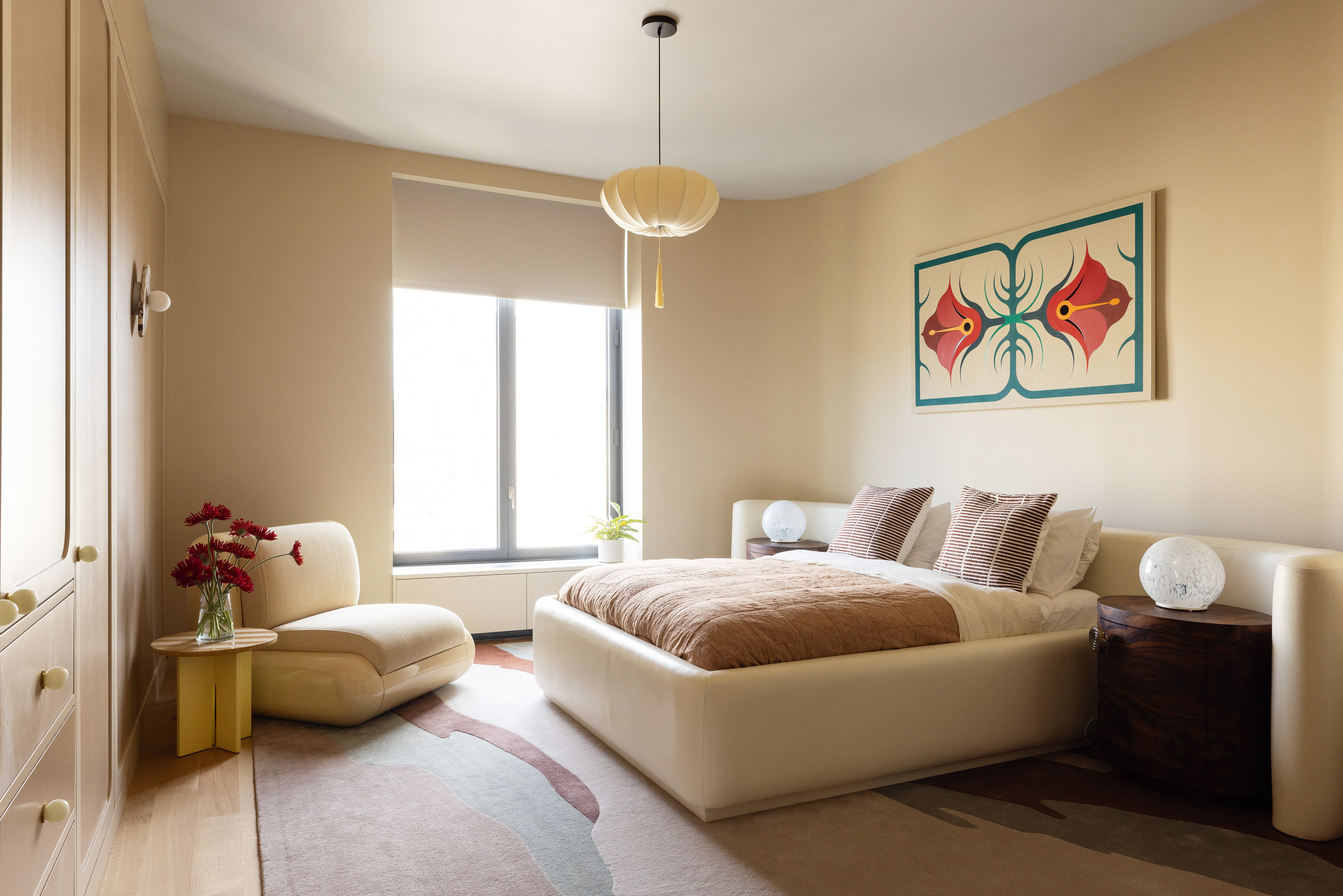 Cream bedroom with cream bed with built-in headboard and dark wood side tables, cream accent chair and pale oak side table