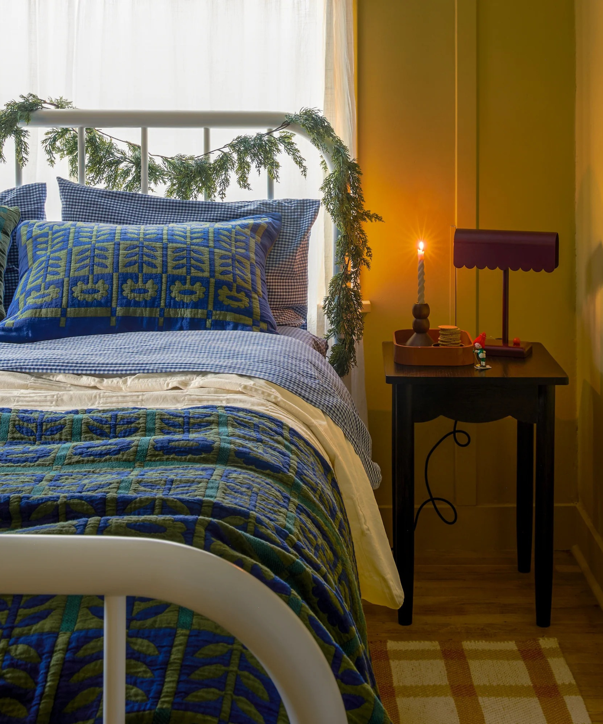 a room with a blue and green floral quilt, a white metal bedframe, a small side table with a lamp on top an a glowing candle
